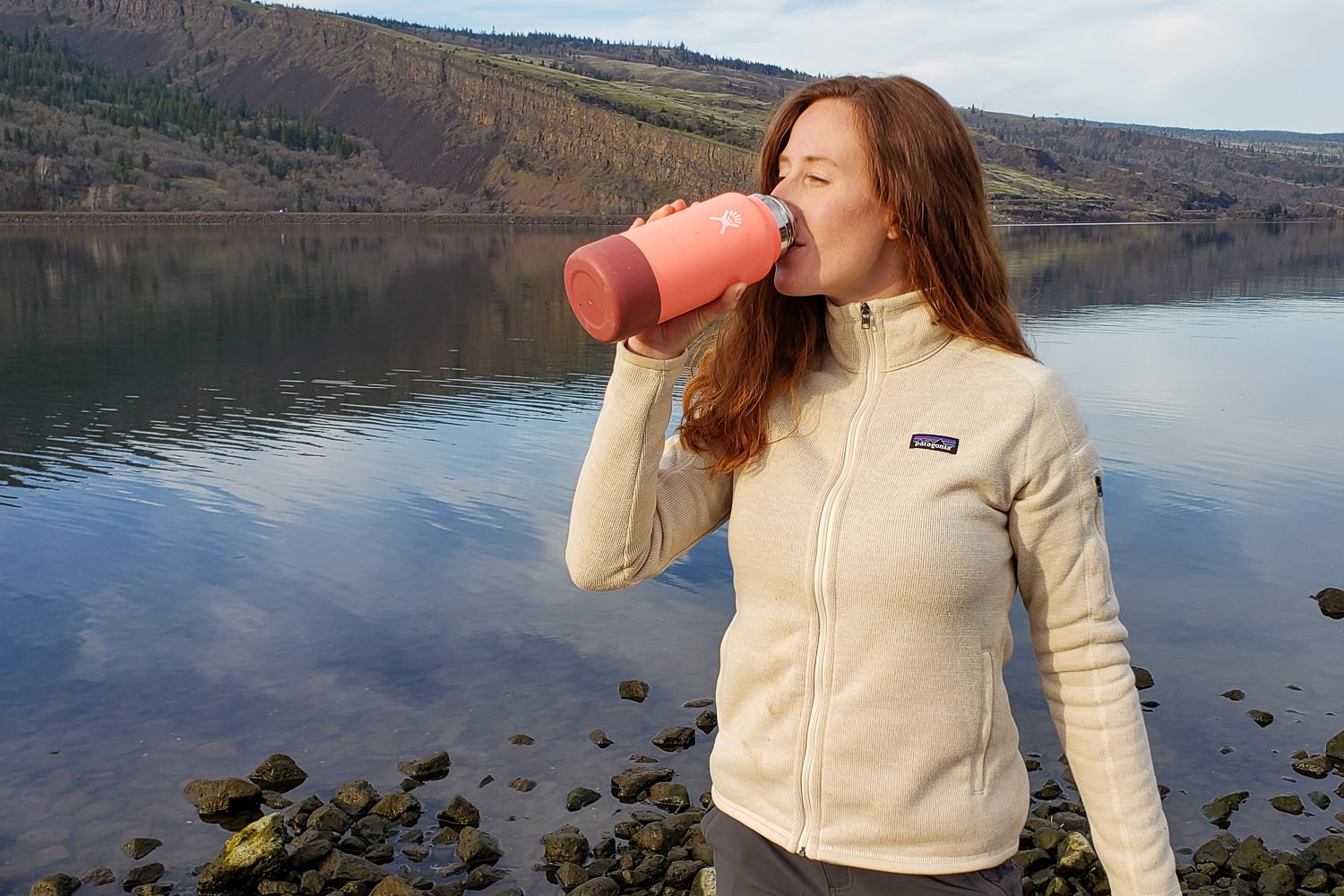 A person drinking from a Hydro Flask with a bottle boot on it in front of a river view