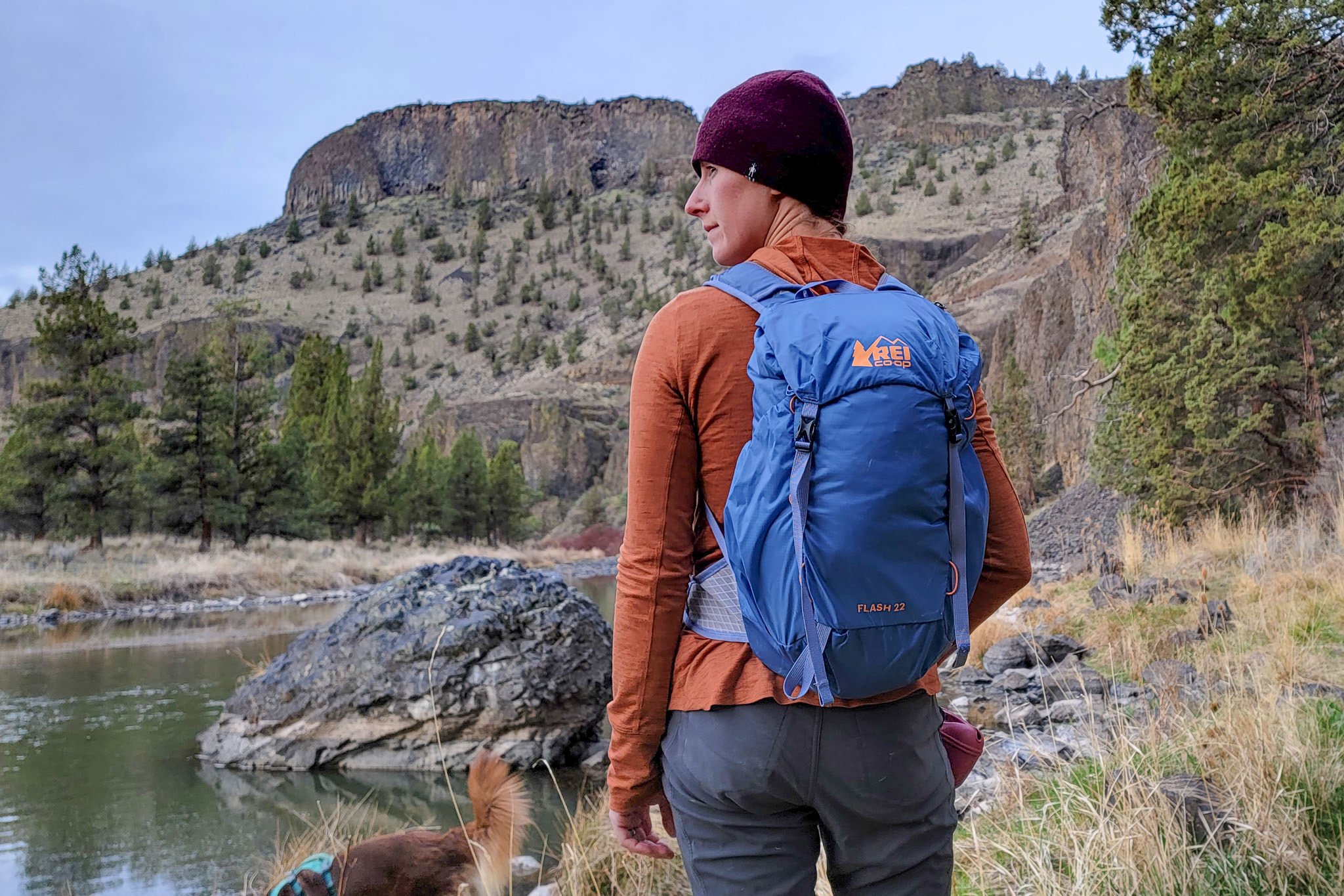 A hiker wearing the REI Flash 22 daypack next to a river with some high desert cliffs in teh background