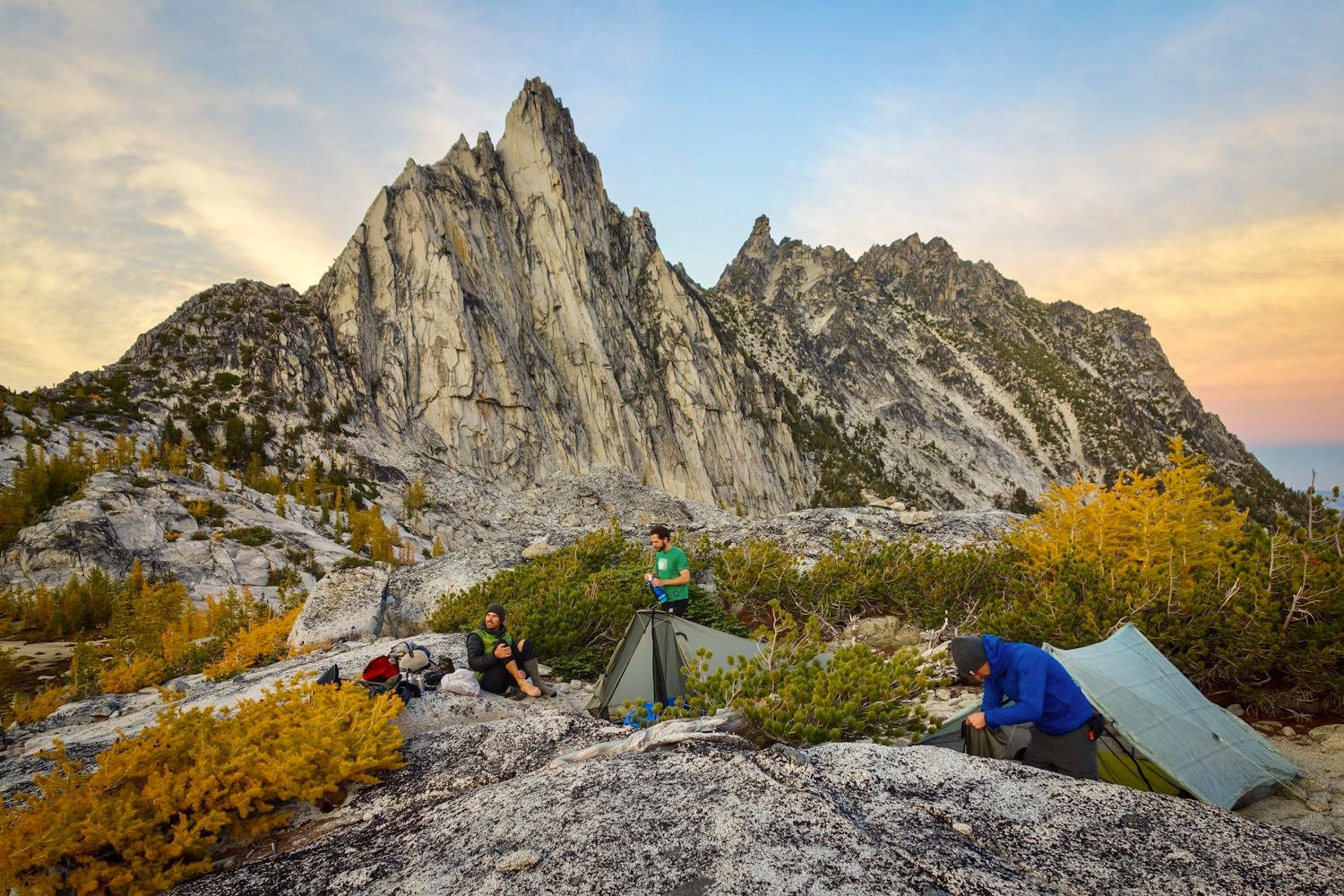 Three hikers and two tents in front of a beautiful mountain at sunset