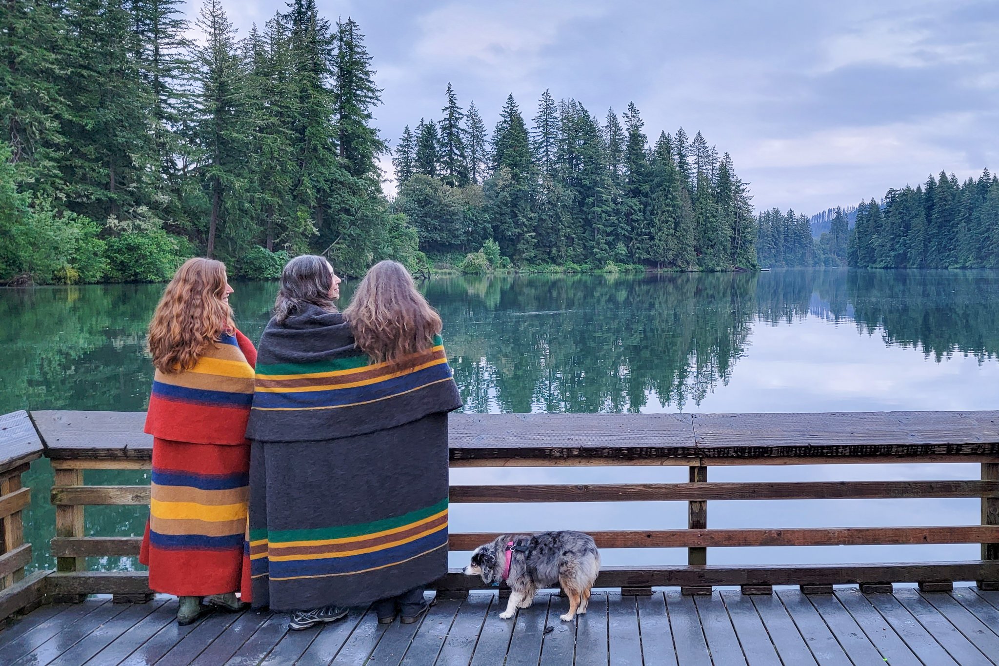 Three women bundled up in the Pendleton Yakima & National Parks Blankets on a dock over a lake