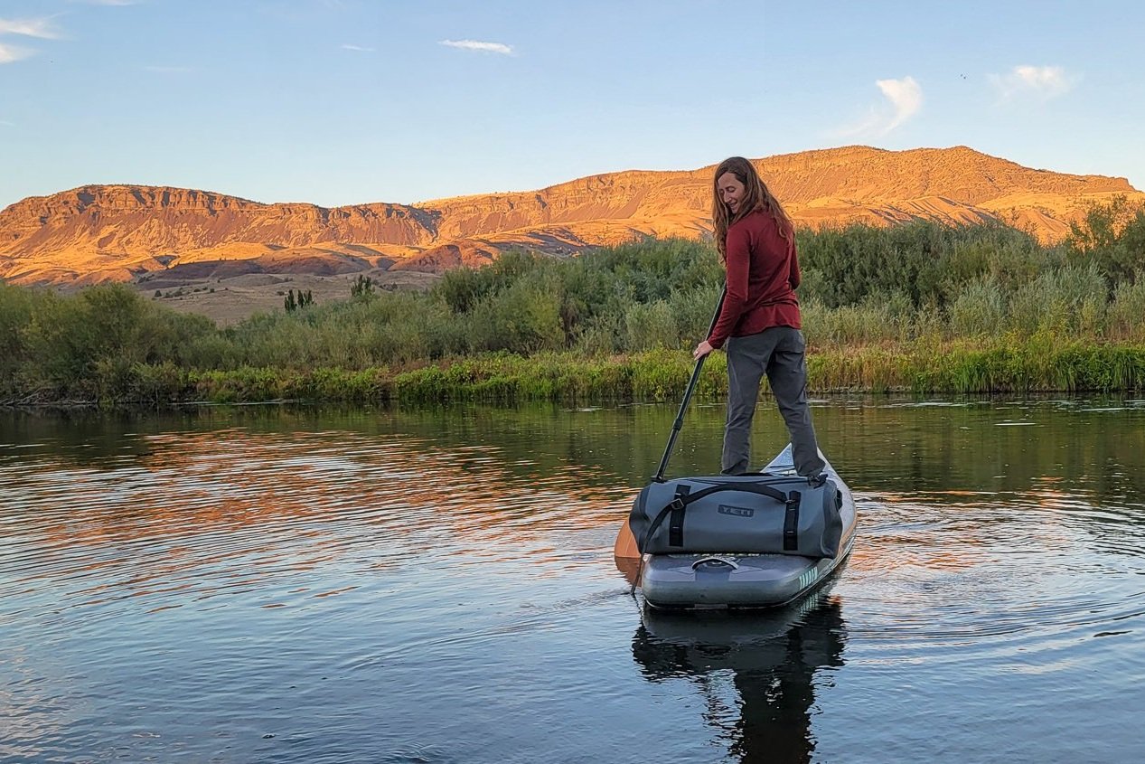 A woman paddleboarding on a river with a mountain in the background - the Yeti Panga 75 duffel is on the back of the paddleboard
