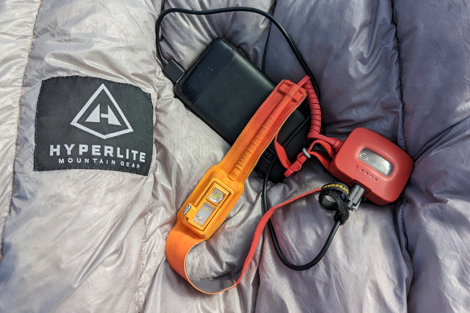 A red Biolite HeadLamp 325 sitting on top of a backpacking quilt. The headlamp is plugged into a power bank to charge