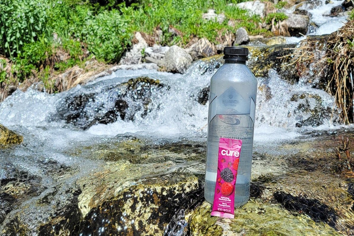 A cure hydration stick pack next to a bottle of water in front of a rushing creek