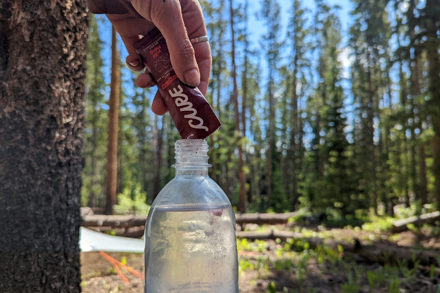 A packet of cure hydration mix being poured into a water bottle in a forest