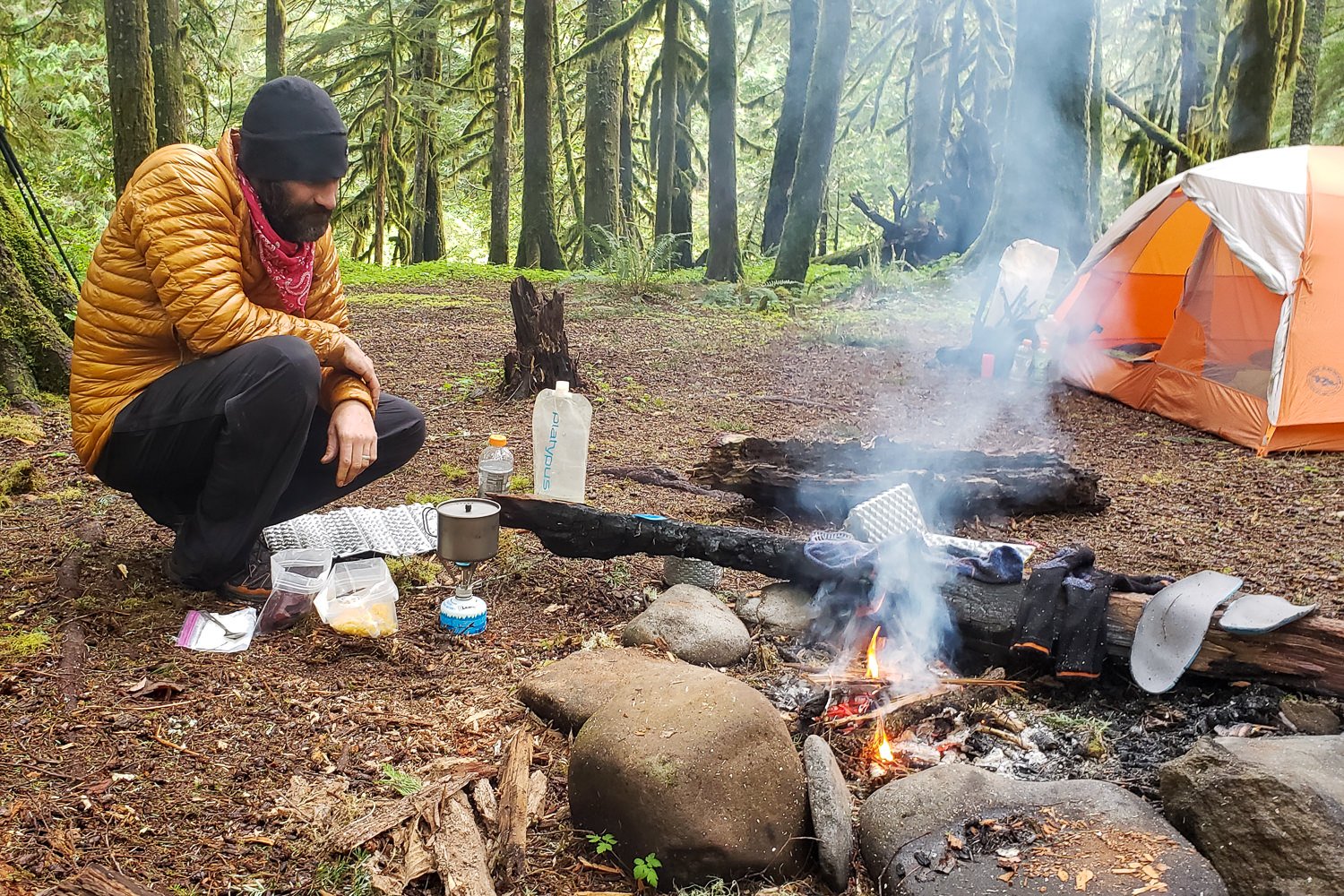 A hiker sitting next to a small fire in a fire ring at a campsite