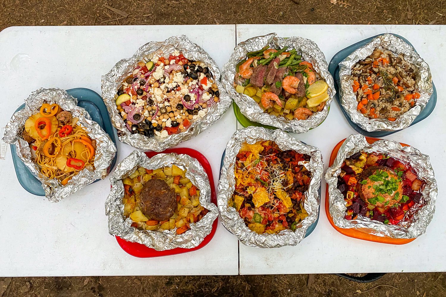 Top-down view of seven different cooked foil packet meals on a camping table