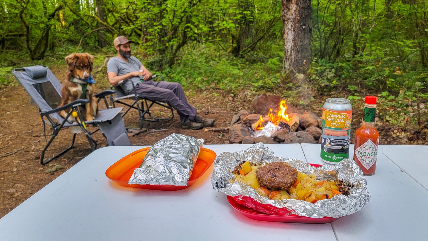 Closeup of a classic foil packet meal with a man and a dog sitting around a campfire in the background