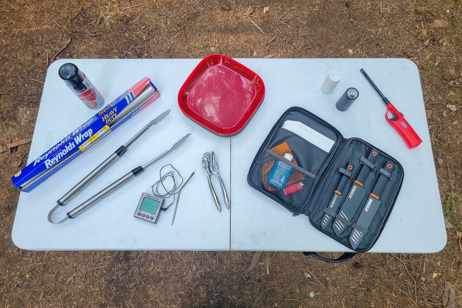 A camping table with foil, non-stick cooking spray, tongs, a meat thermometer, can opener, plates, camping knife set, salt and pepper, and a lighter laid out