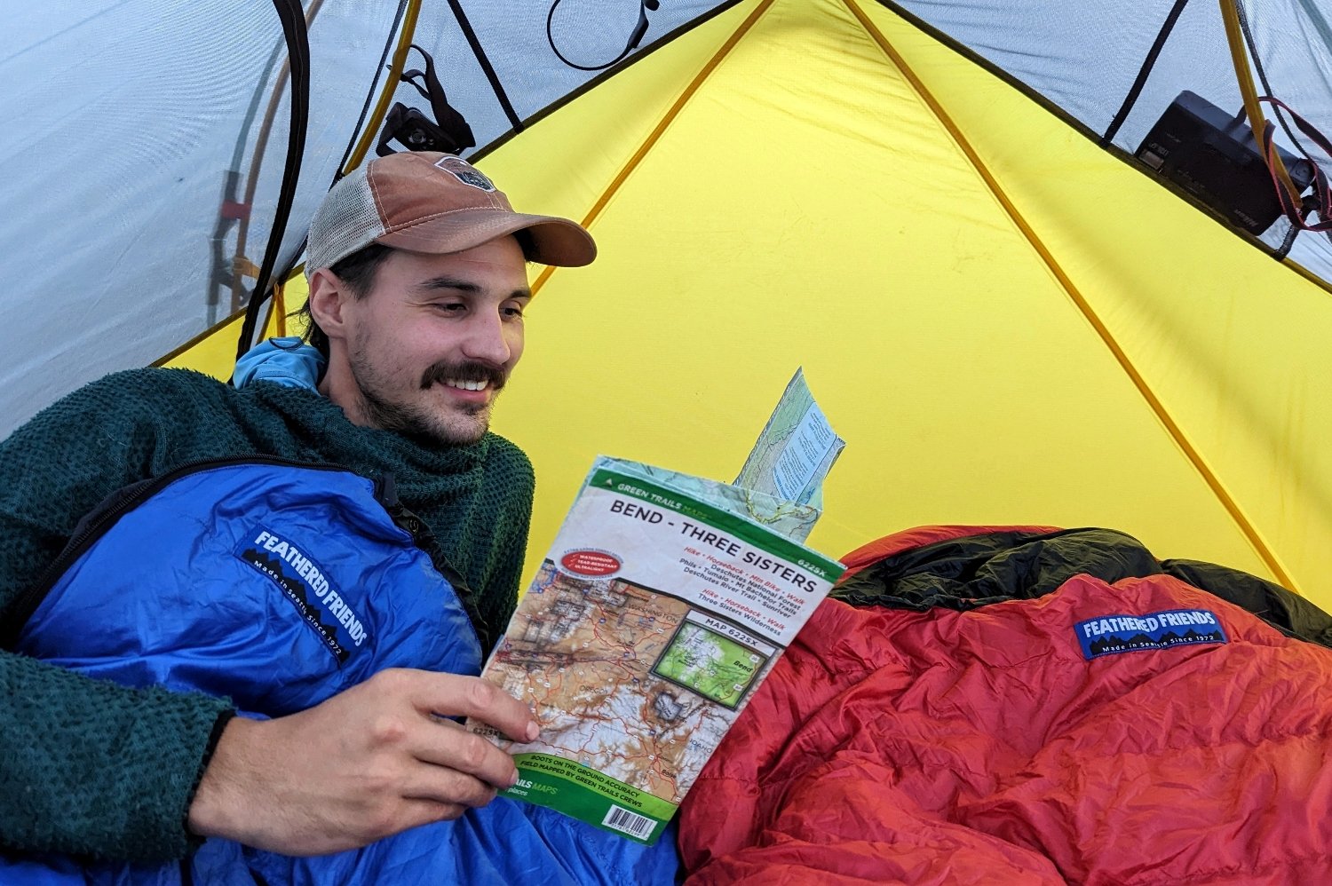 A hiker in a tent bundled up in a Feathered Friend Swallow YF 20 sleeping bag. He's studying a map of the Three Sisters Wilderness