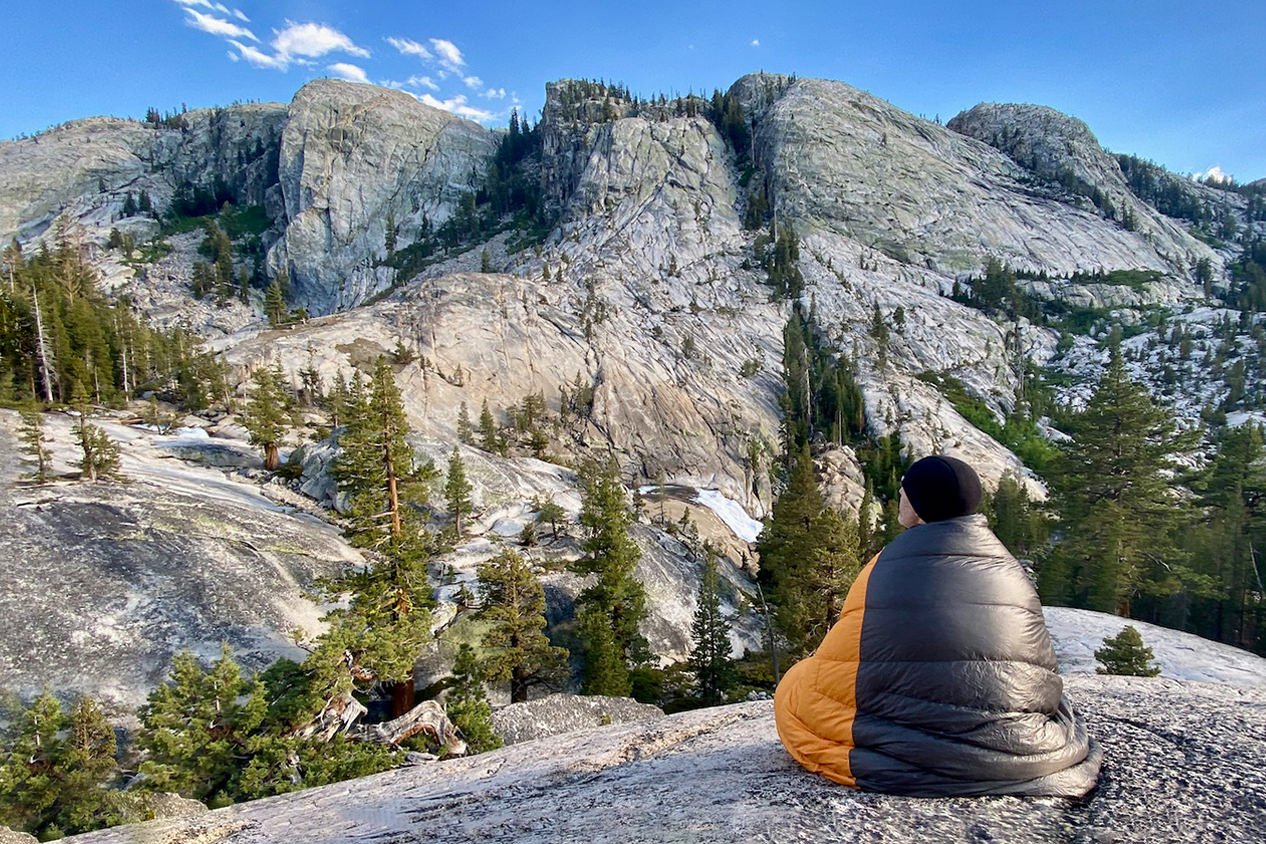 A hiker wrapped up in an Enlightened Equipment Revelation Quilt looking at a mountain view