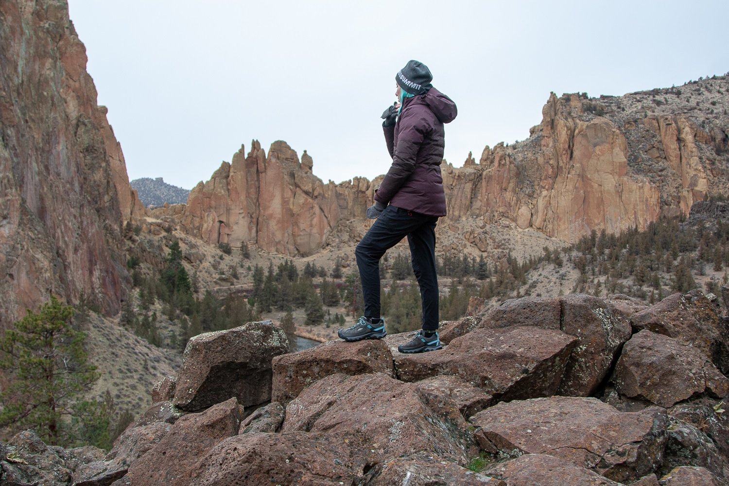 A hiker wearing the REI Stormhenge jacket on a rocky outcrop with somejagged peaks in the background. The sky is gray