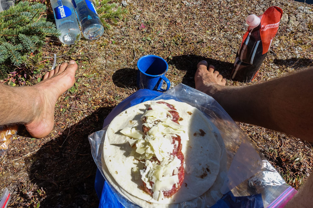 Looking down at a tortilla with salami, cheese, and condiments between a hikers feet.