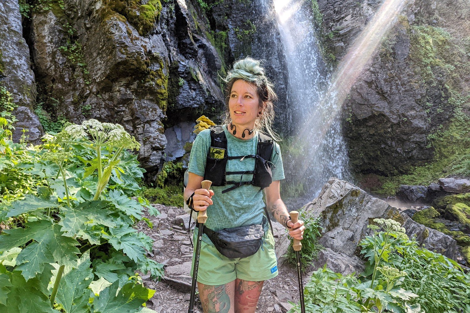 A smiling hiker wearing a Hyperlite Mountain Gear Versa fanny pack with a big waterfall in the background. There are bright green plants around and a ray of sunlight is shining through the water cascading down