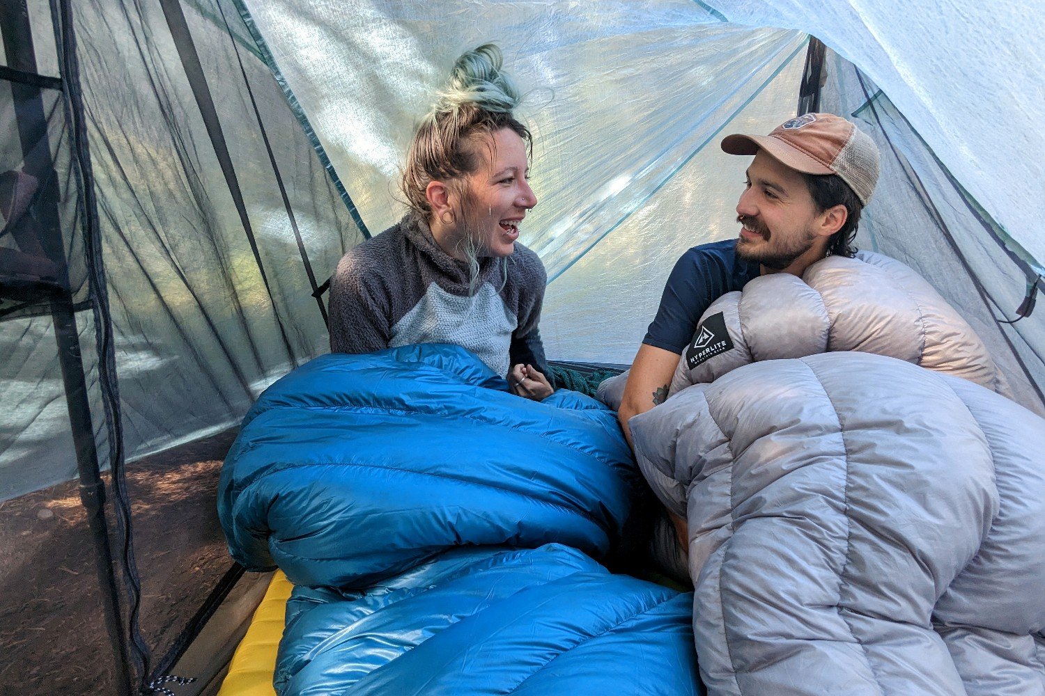 Two hikers laughing with each other wrapped up in backpacking quilts inside a backpacking tent