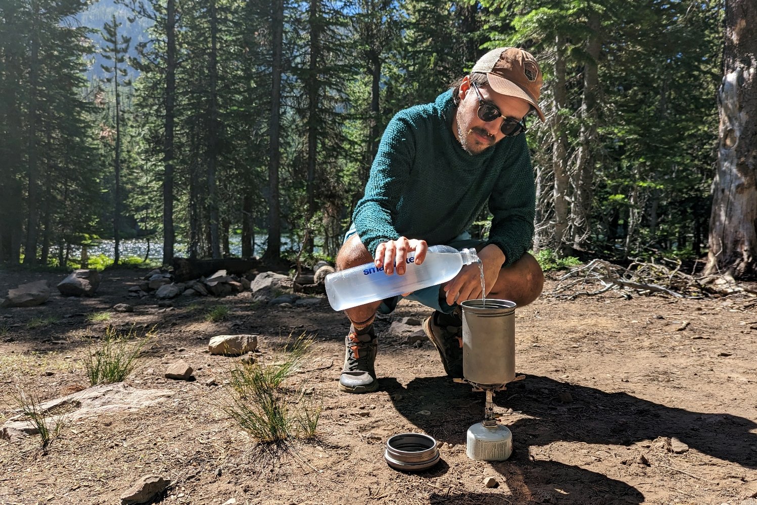 A hiker squatting down to pour water from a Smartwater Bottle in a Vargo Bot that's sitting on top of a backpacking stove. There are pine trees and a lake in the background